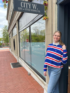 A person standing on a sidewalk in front of a store window displaying a striped long-sleeve rugby shirt in red, white, and blue.